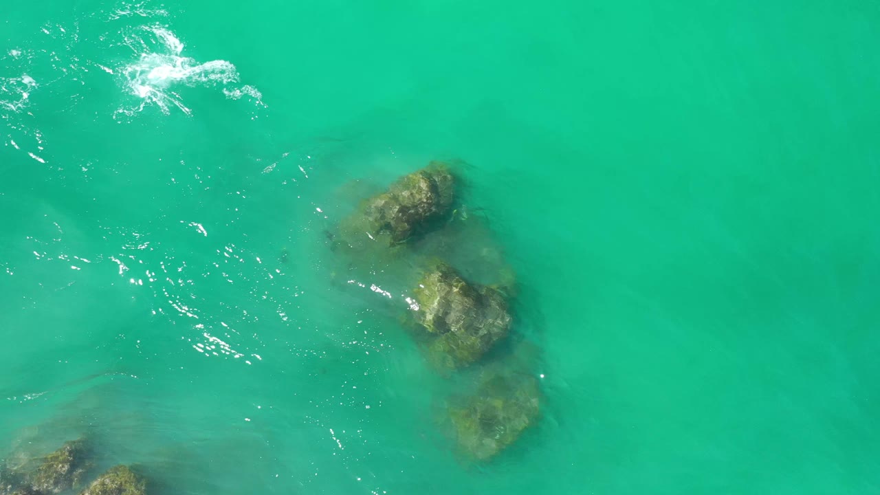 Overhead Los Cabos water frame showing submerged rock shapes near the right side, scattered white foam near the top, and no visible people.