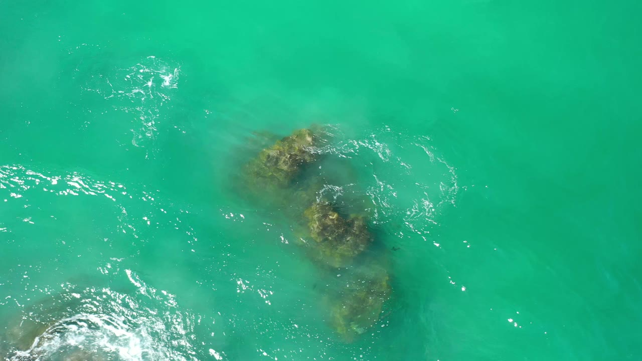 Overhead Los Cabos sea view with dark submerged formations centered lower in frame, bright ripples to the left, and no visible people.