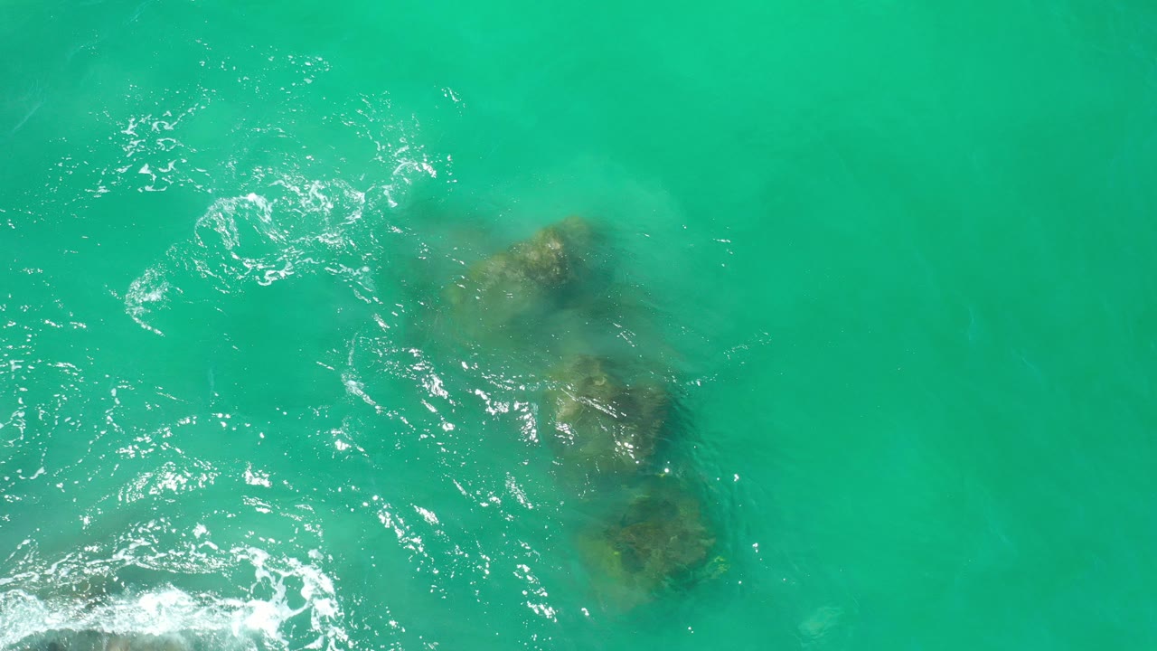 Overhead view in Los Cabos, Mexico of green ocean water with a cluster of submerged rocks near the lower center, white ripples at left, and no visible people.
