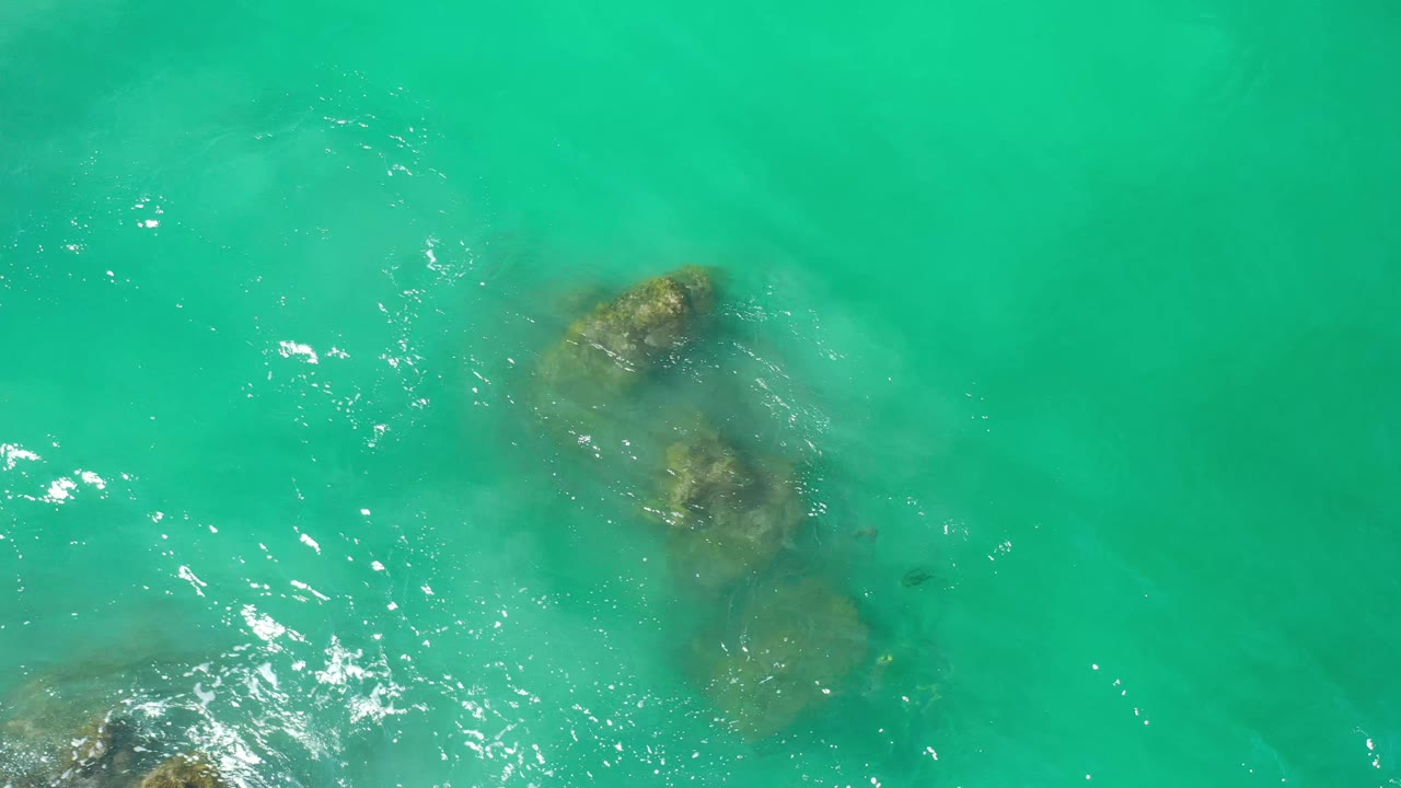Overhead Los Cabos water view with rocks just below the surface, scattered bright reflections, and no visible people.