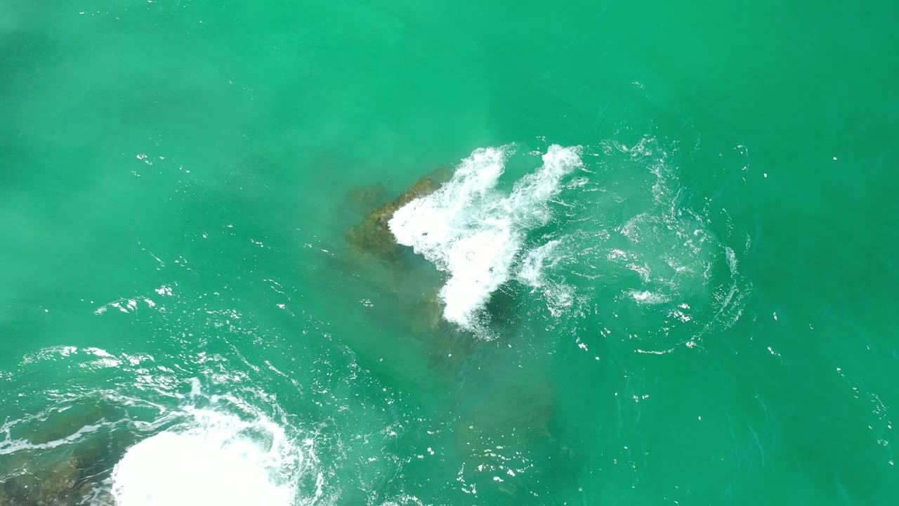 Overhead view of turquoise water in Los Cabos, Mexico, with white foam breaking over dark submerged rocks and no visible people.