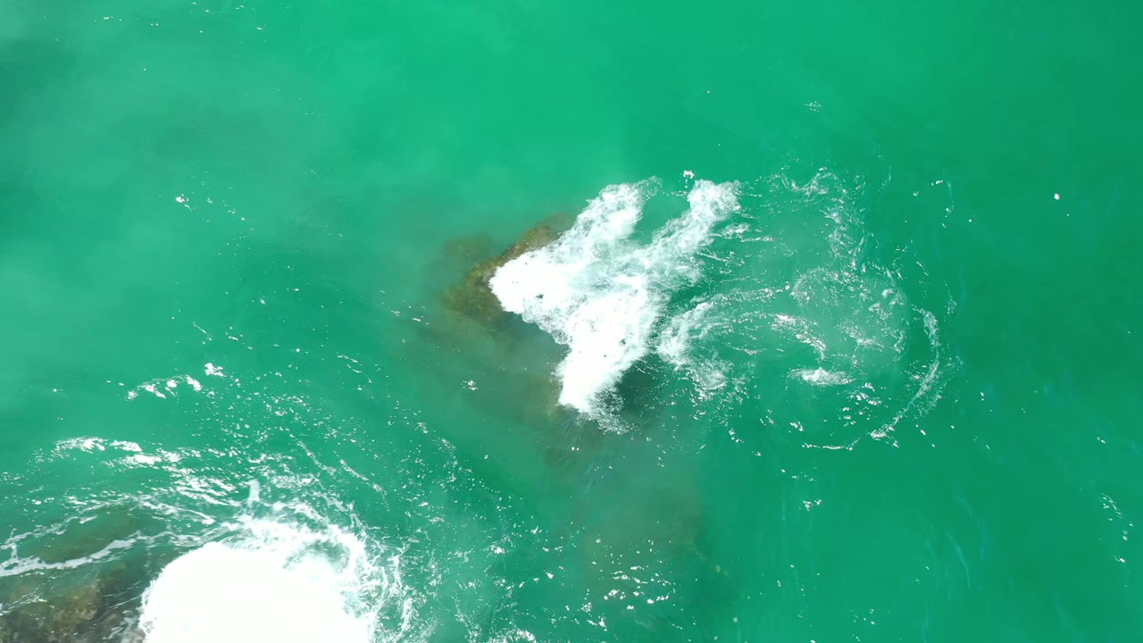 Overhead Los Cabos water view with foam trails sliding across a green-blue surface and a dark rock shape under the water, with no visible people.
