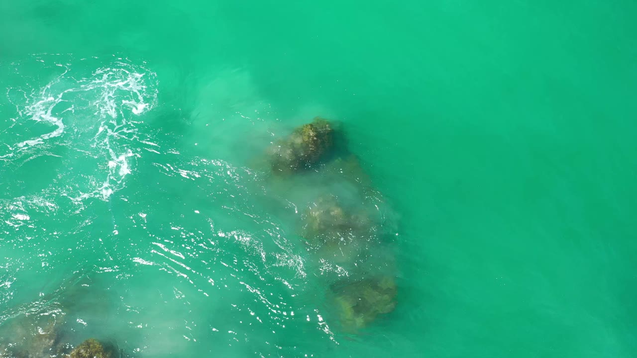 Overhead Los Cabos frame showing green-turquoise water, a cluster of submerged rocks to the right, ripples across the center, and no visible people.