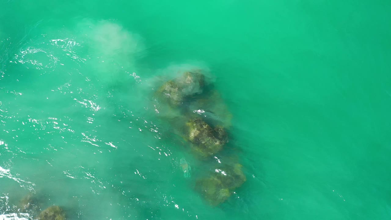 Overhead Los Cabos water view with clustered submerged rocks near the right side, foam drifting above them, and no visible people.