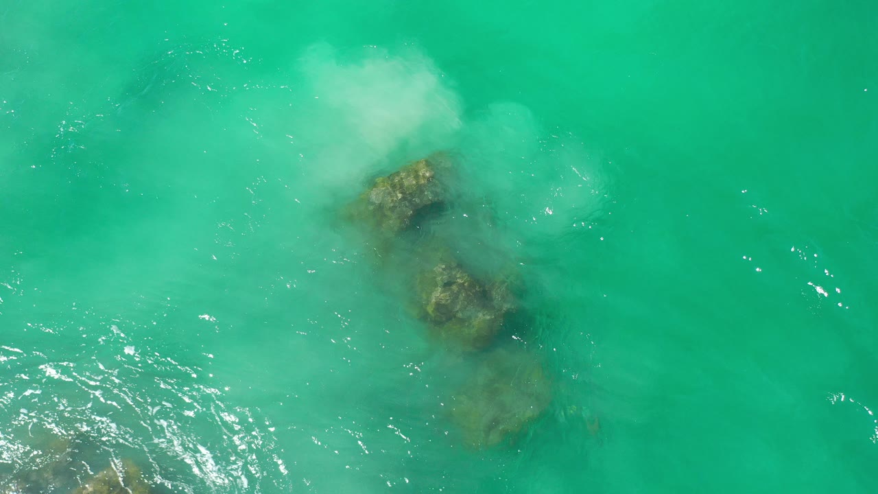 Overhead view of turquoise water in Los Cabos, Mexico, with submerged rocks below a patch of white foam and no visible people.