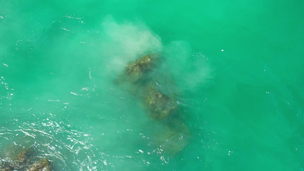 Blue-green ocean surface with rocks under water and a foam cloud at top right.
