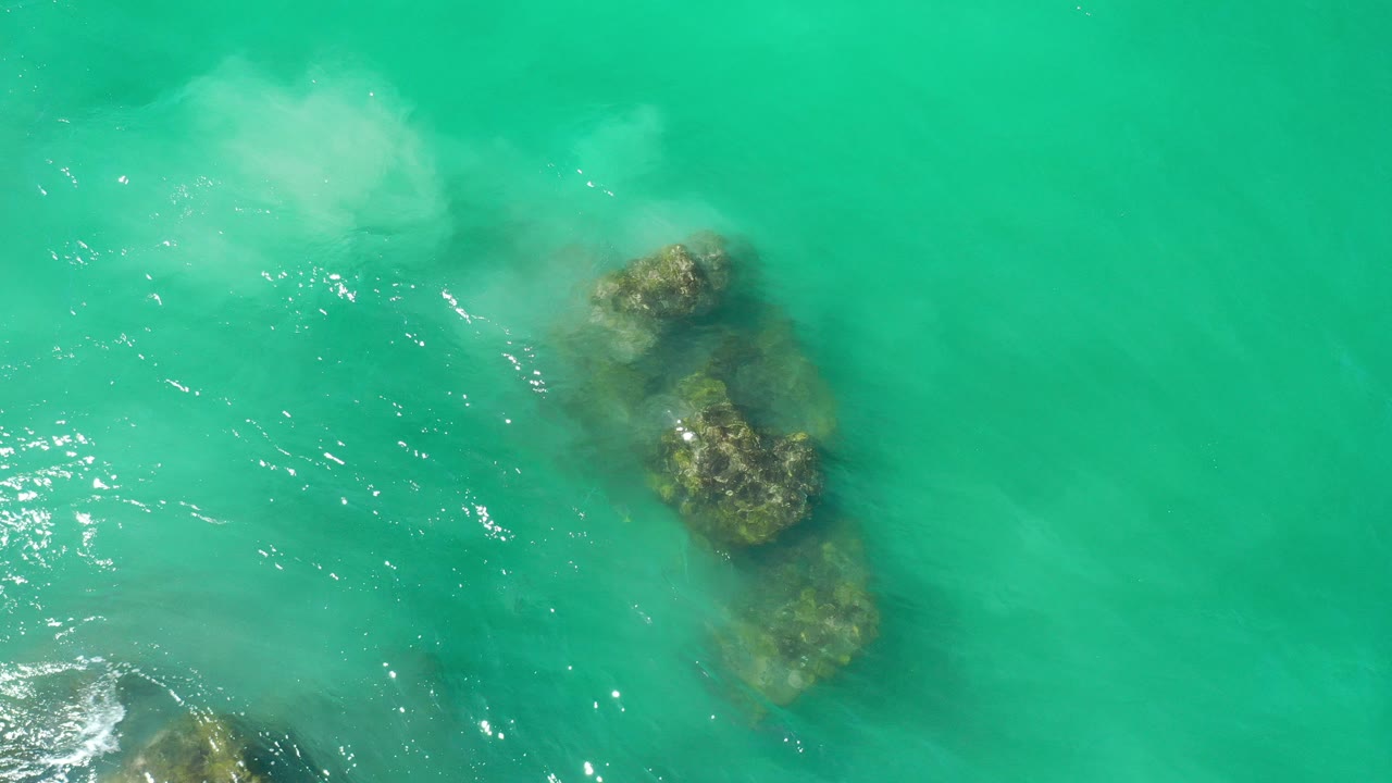 Overhead view of greenish-blue water in Los Cabos, Mexico, with clustered submerged rocks near the center and pale foam near the top, with no visible people.