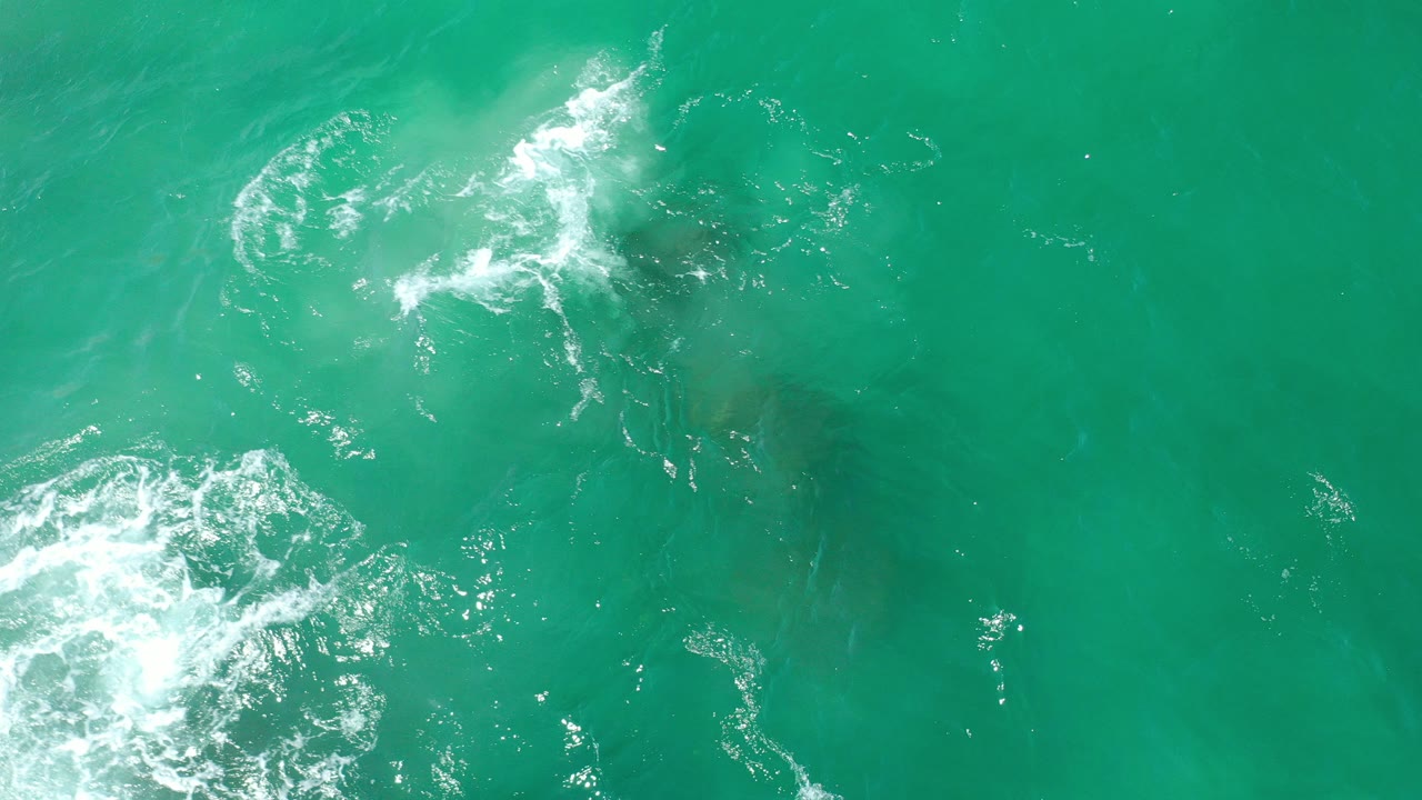Overhead view of turquoise water in Los Cabos, Mexico, with white foam near the left edge, darker underwater rock shapes below the surface, and no visible people.