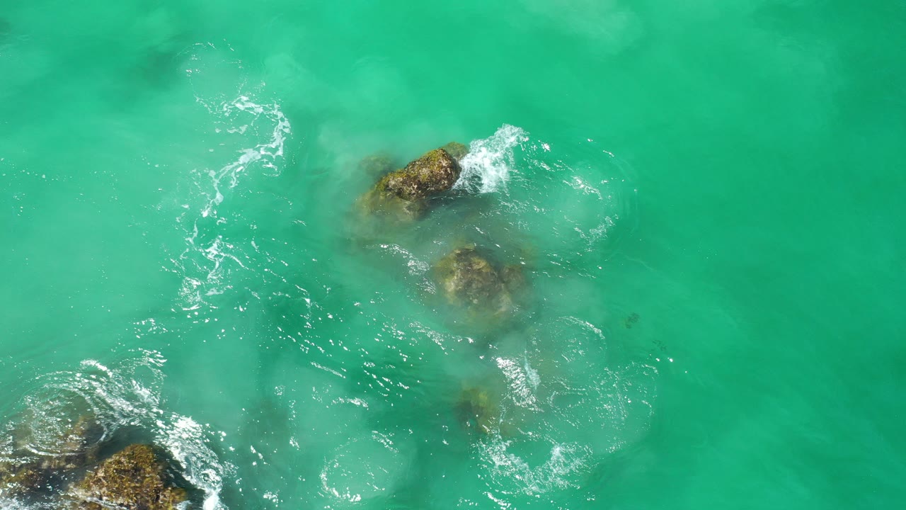 Overhead view of turquoise water in Los Cabos, Mexico, swirling around submerged brown rocks with small white foam streaks and no visible people.