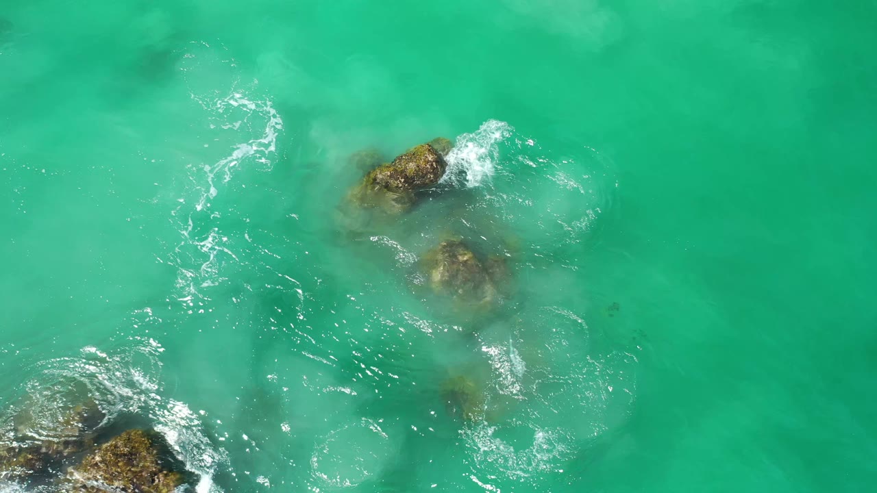 Turquoise Los Cabos water seen from above, with several submerged rocks clustered near the lower and upper frame and no visible people.