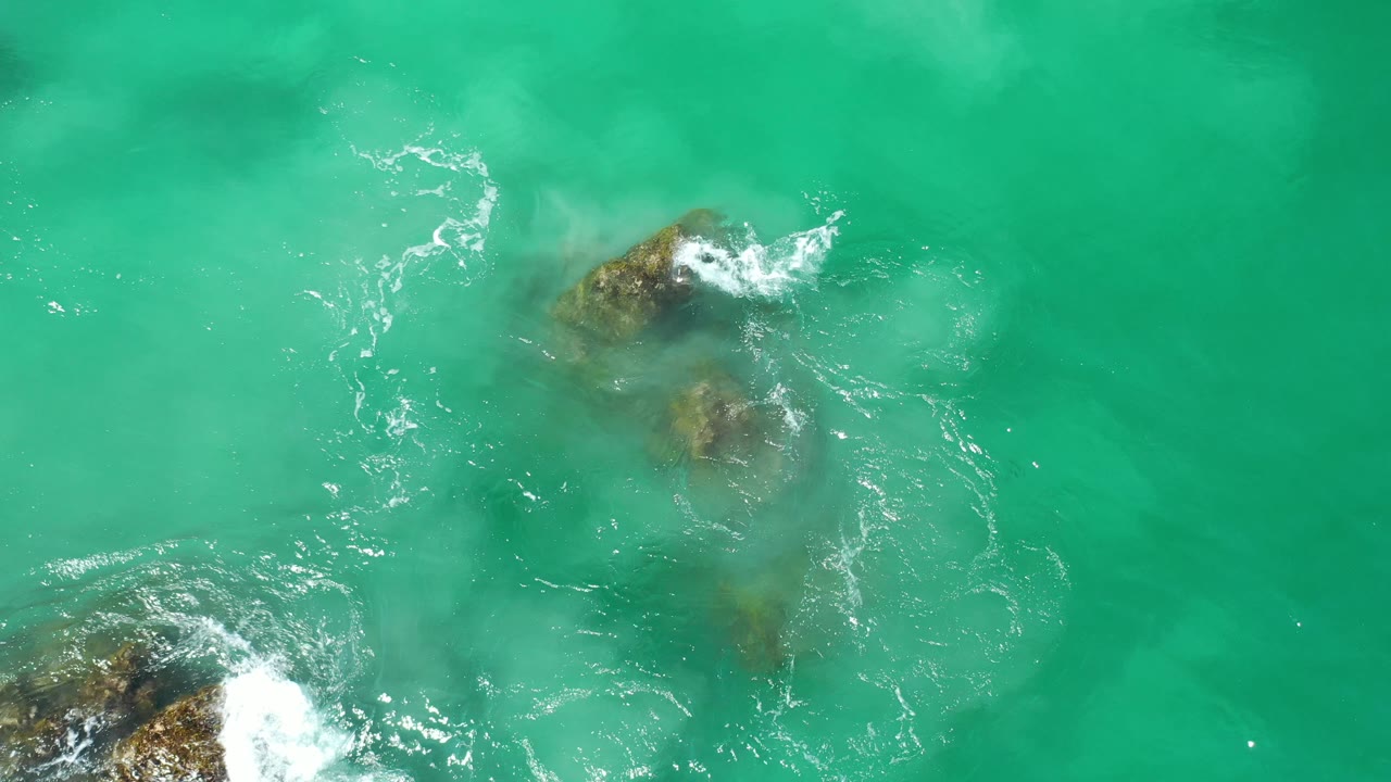 White wave foam breaks over rocks in clear green-blue water.