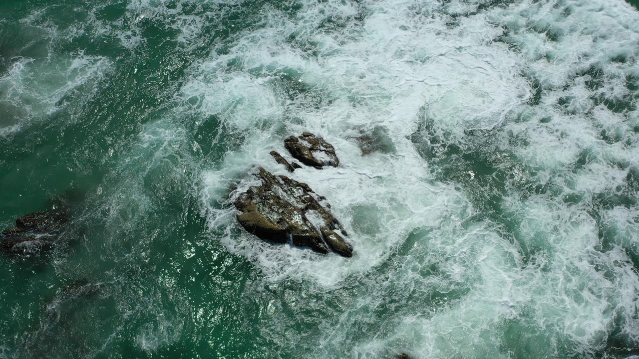 Overhead view of waves breaking around jagged rocks in green ocean water in Los Cabos, Mexico, with no visible people.