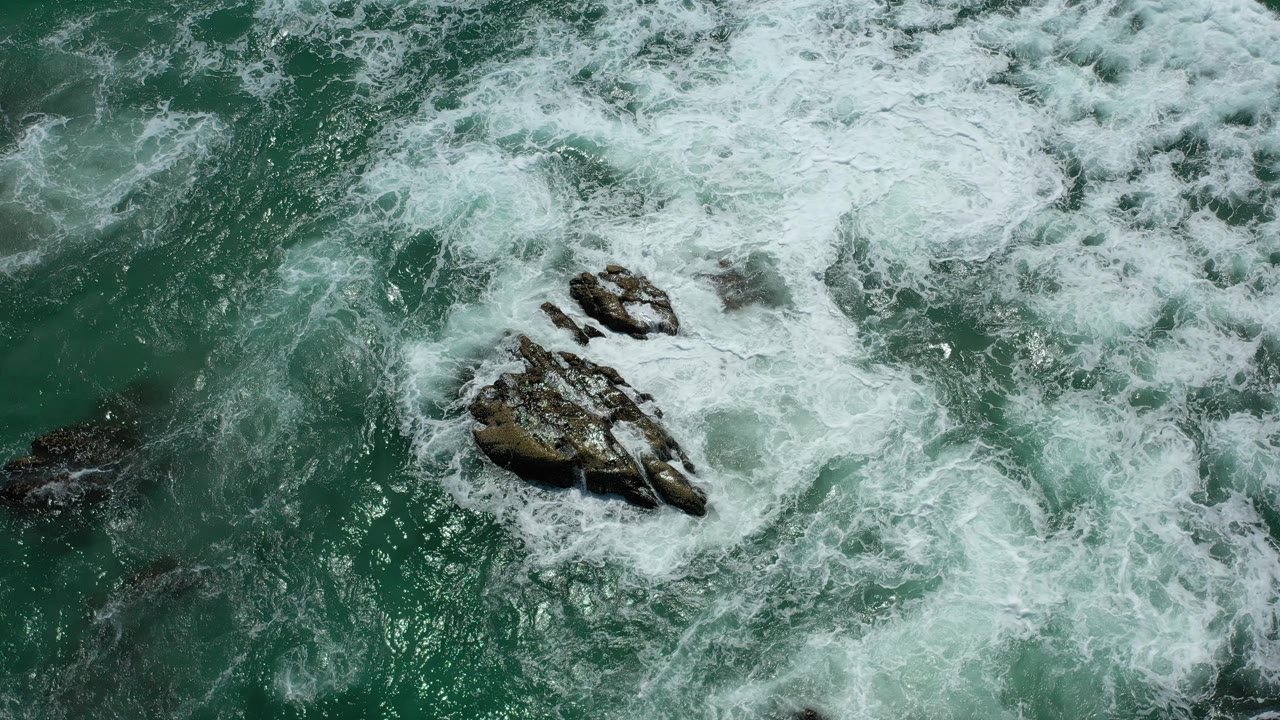White sea foam spreads over and around the rock cluster in green water in Los Cabos, Mexico, with no visible people.