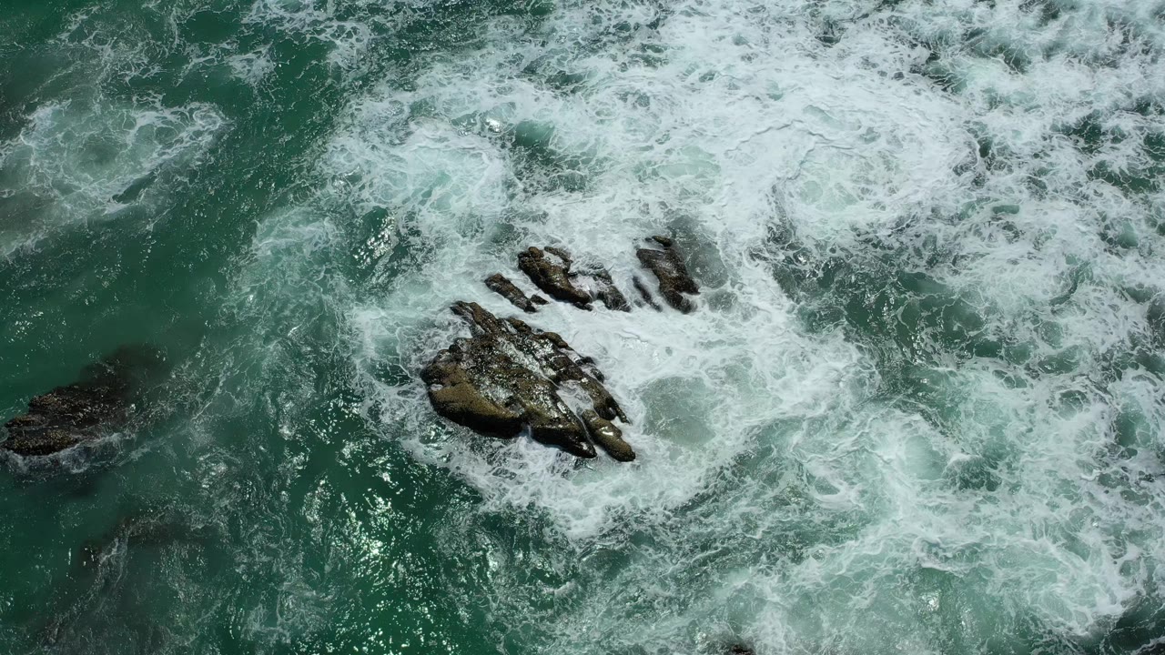 Surf curls around a rock formation seen from above.