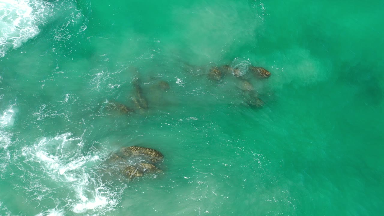 Overhead view in Los Cabos, Mexico of green sea water moving around several submerged rocks, with white foam entering from the left and no visible people.