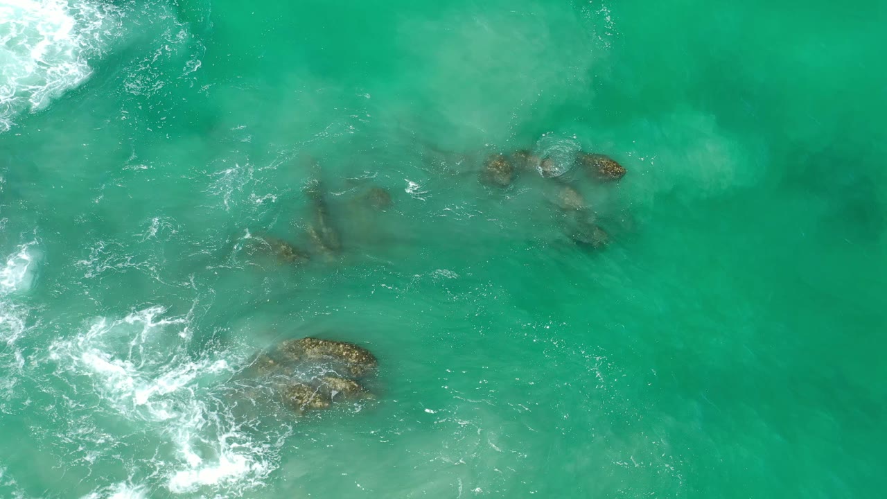 Overhead Los Cabos frame with turquoise-green water, a larger exposed rock near the lower center, smaller rocks near the middle, and no visible people.