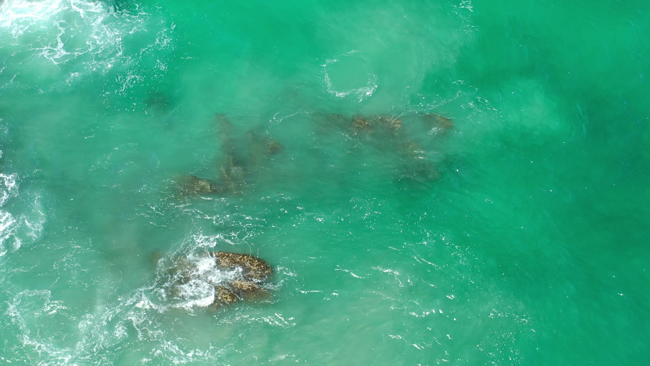 Overhead view of green seawater and white foam breaking around several rocks in Los Cabos, Mexico, with no visible people.