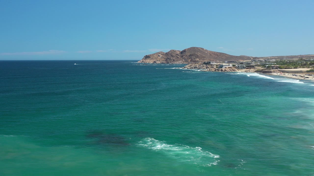 Aerial view over the Los Cabos, Mexico coast with turquoise ocean in the foreground, a sandy beach and low buildings to the right, rocky hills beyond, and no visible people.