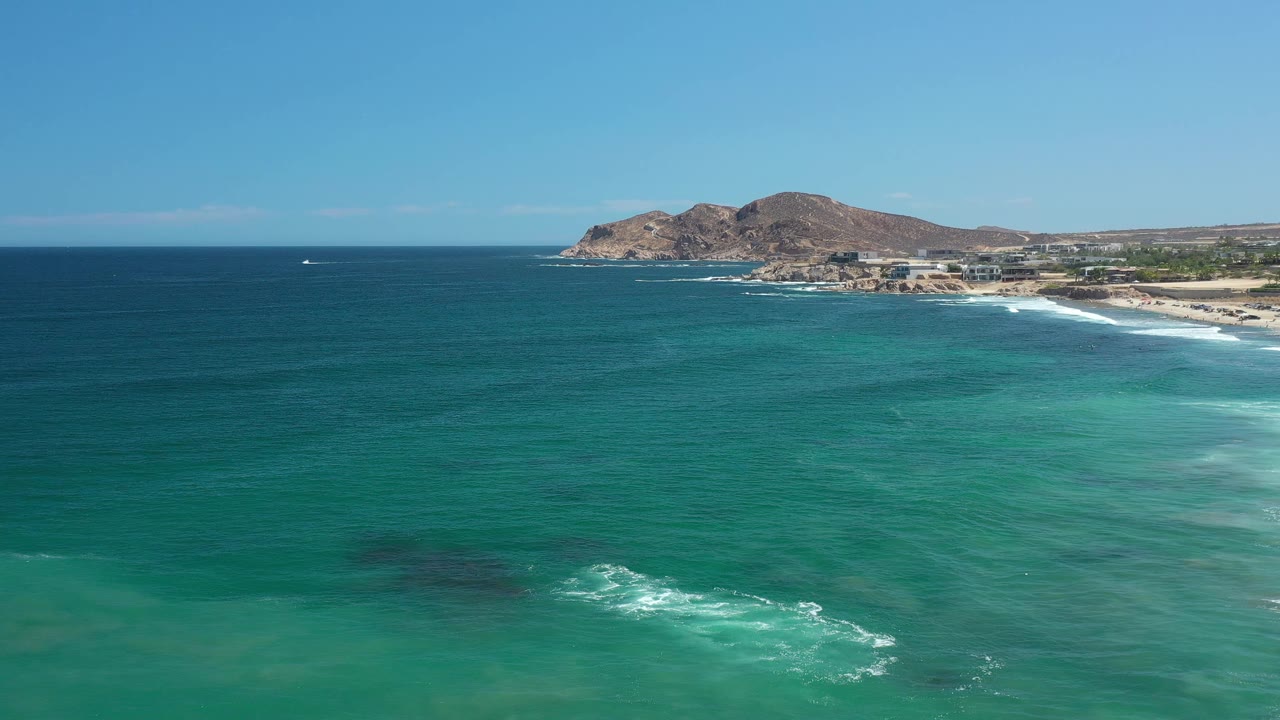 Wide aerial frame of the Los Cabos coastline with clear blue water across most of the image, surf breaking near shore, a small white boat far offshore, and no visible people.