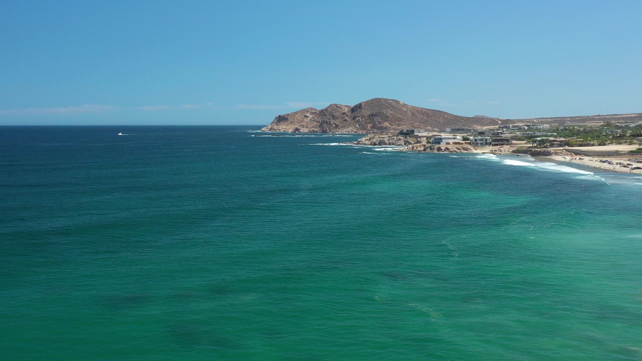 Aerial frame over the Los Cabos coast with turquoise water in the foreground, white surf along a sandy beach, rocky points beyond, and no visible people.