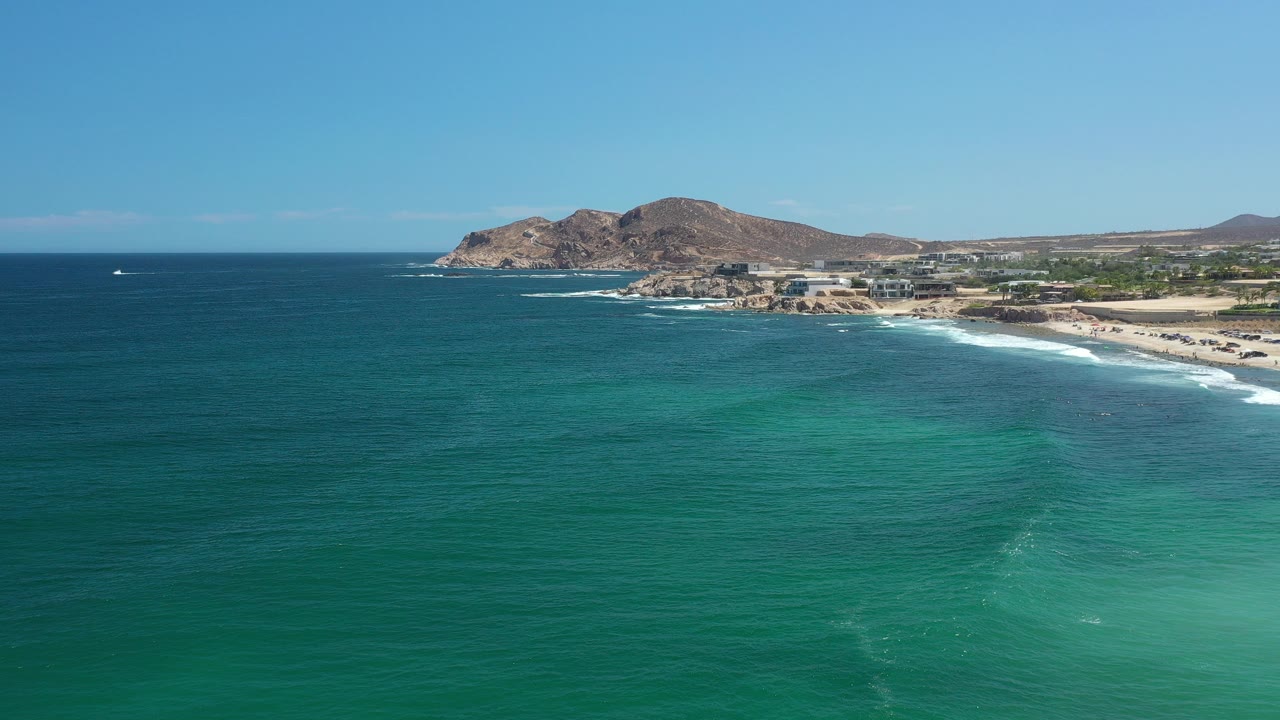 Wide aerial frame over the Los Cabos coast, with turquoise ocean in the foreground, white surf along the beach, rocky hills beyond the shore, and no visible people.
