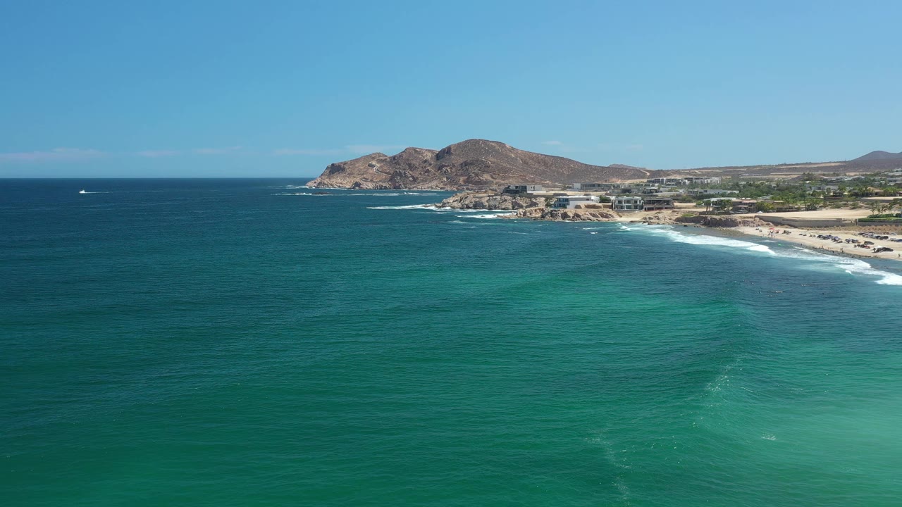 Los Cabos aerial view with darker blue water near the horizon, teal shallows below, a thin surf line on the beach, coastal buildings, and no visible people.