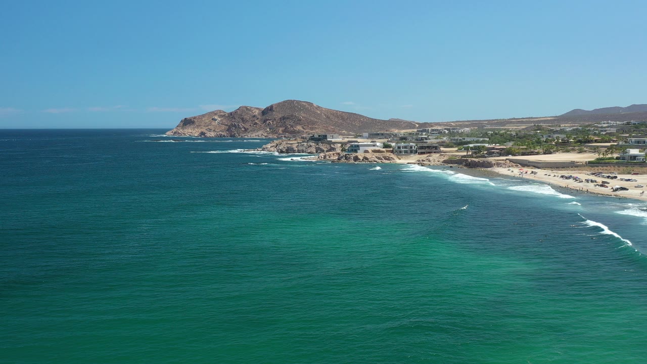 Aerial view of turquoise ocean and a sandy shoreline with rocky hills beyond.