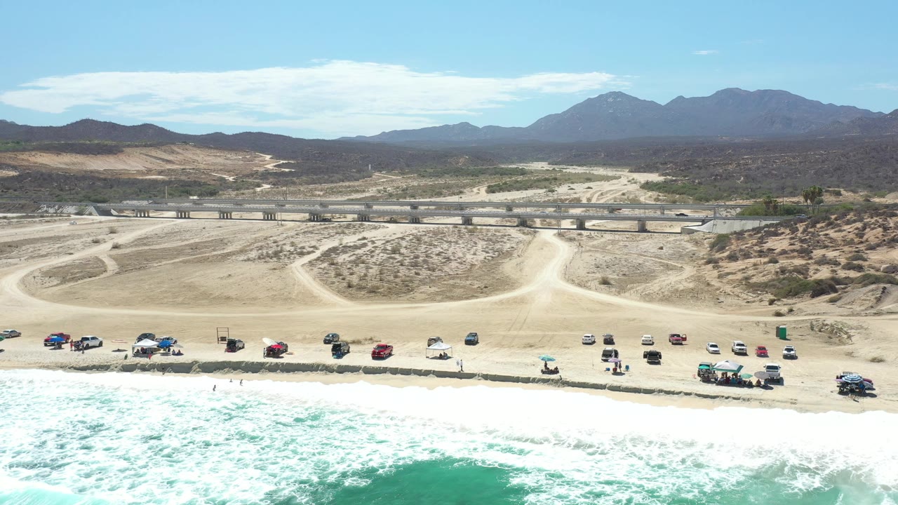 Opening aerial view in Los Cabos, Mexico, with white surf at the bottom edge, parked vehicles along the beach, pale dirt tracks inland, and mountains on the horizon, with no clearly visible people.
