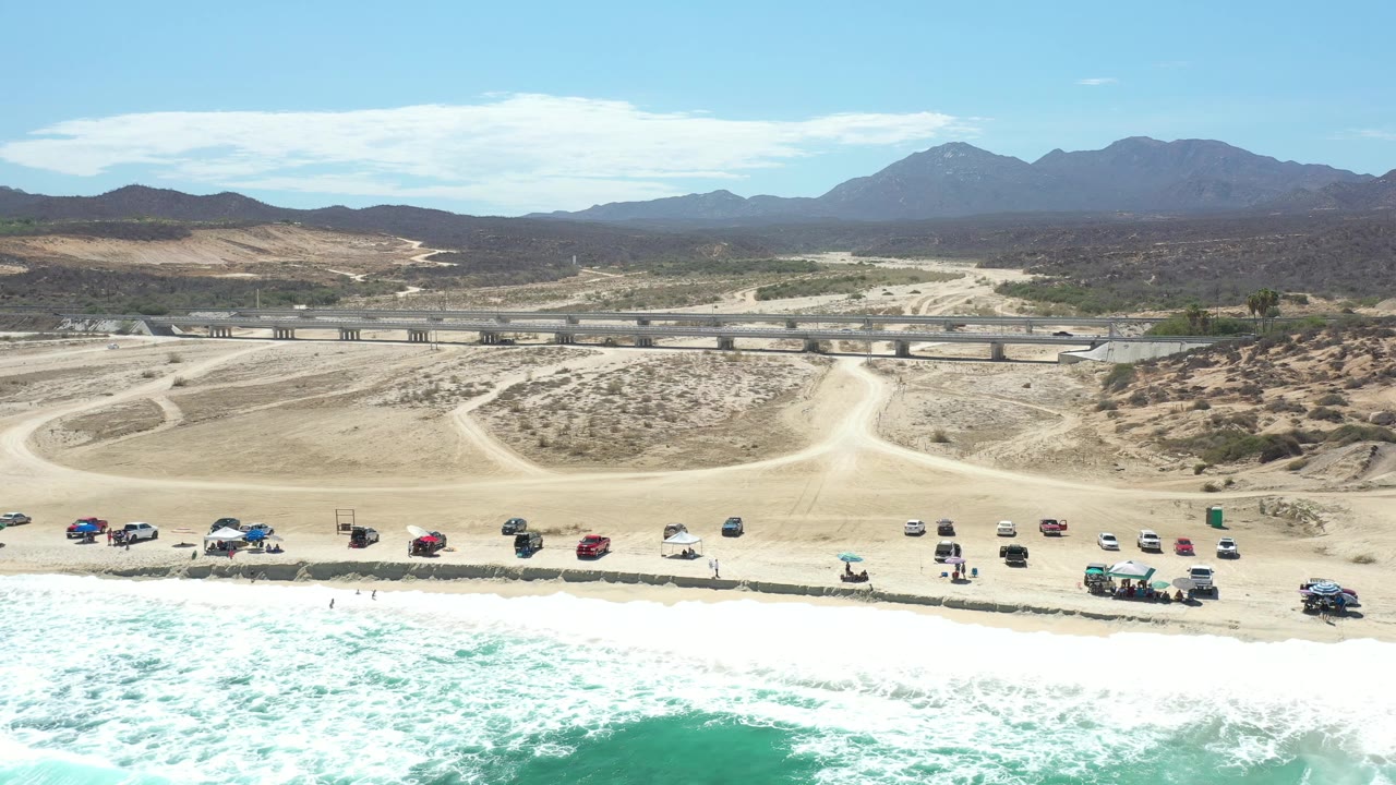 Los Cabos aerial frame showing surf across the lower edge, a line of small parked vehicles on sand, winding dirt road access, scrubby desert ground, and a dark mountain range beyond, with no clearly visible people.