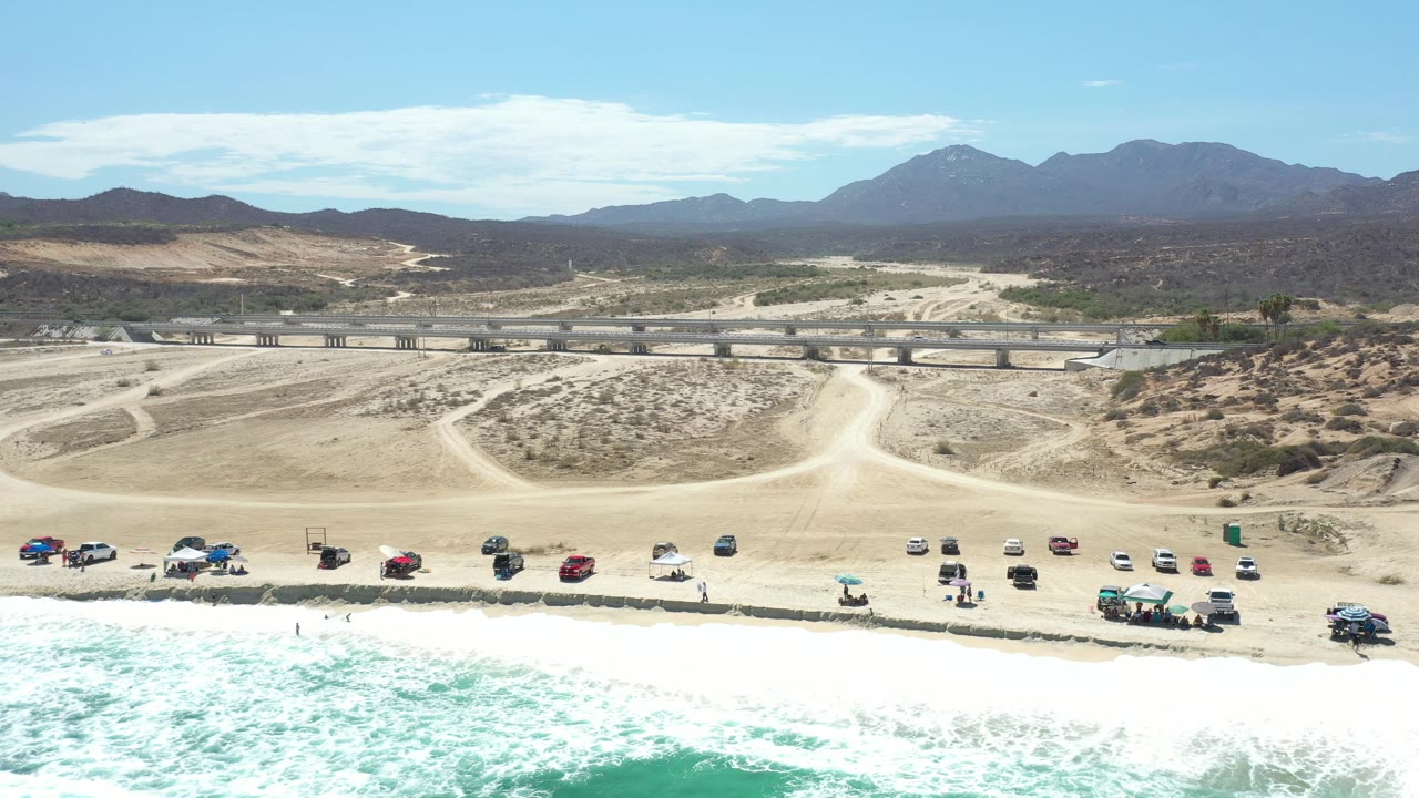Drone-height view of beach parking, surf, inland tracks, bridge, desert, and mountains.