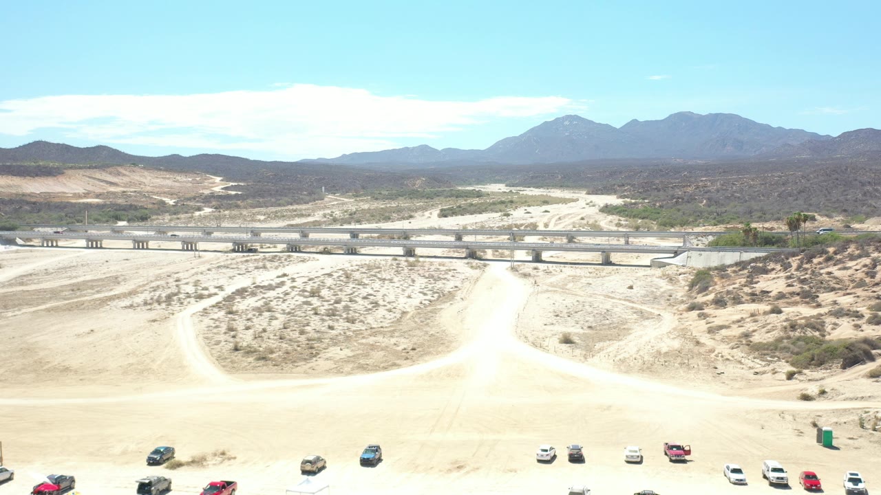 Wide aerial view of desert trails, highway overpass, parked vehicles, and mountains