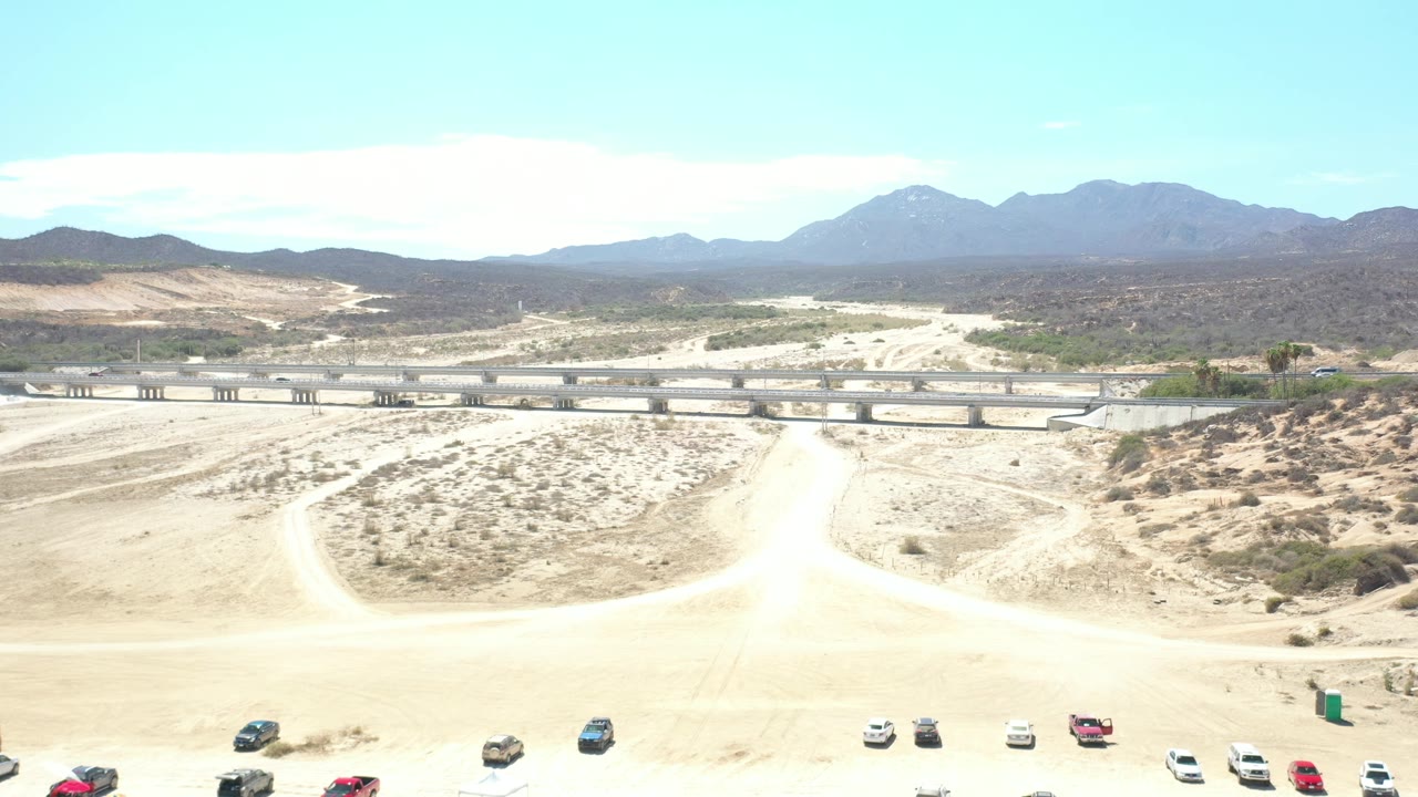 Los Cabos aerial frame with the concrete overpass crossing the center, curved dirt roads in the sandy foreground, low brush, hazy mountains, bright blue sky, and no visible people.