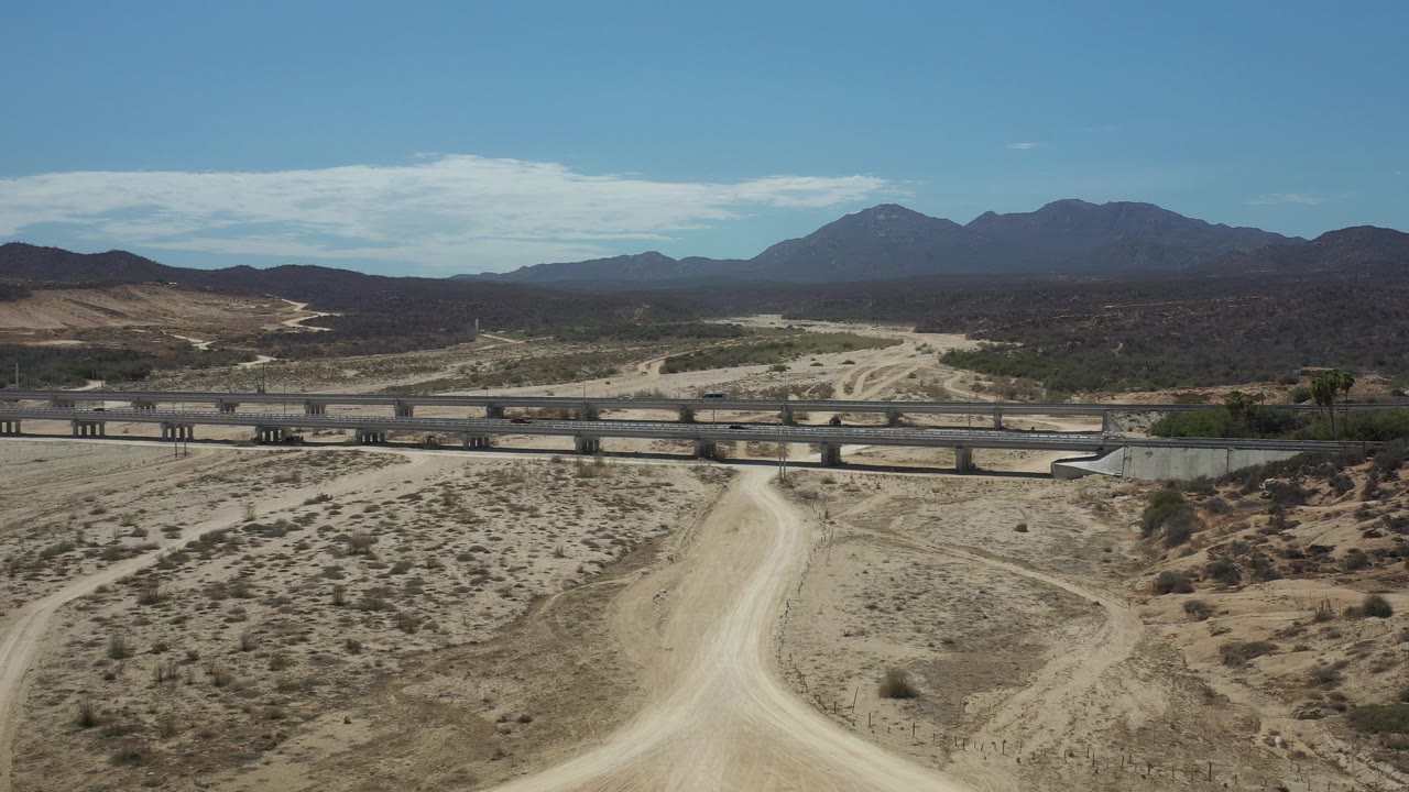 Wide aerial frame in Los Cabos showing a pale dirt road leading toward a highway bridge, sparse desert scrub, distant mountains, blue sky, and no visible people.