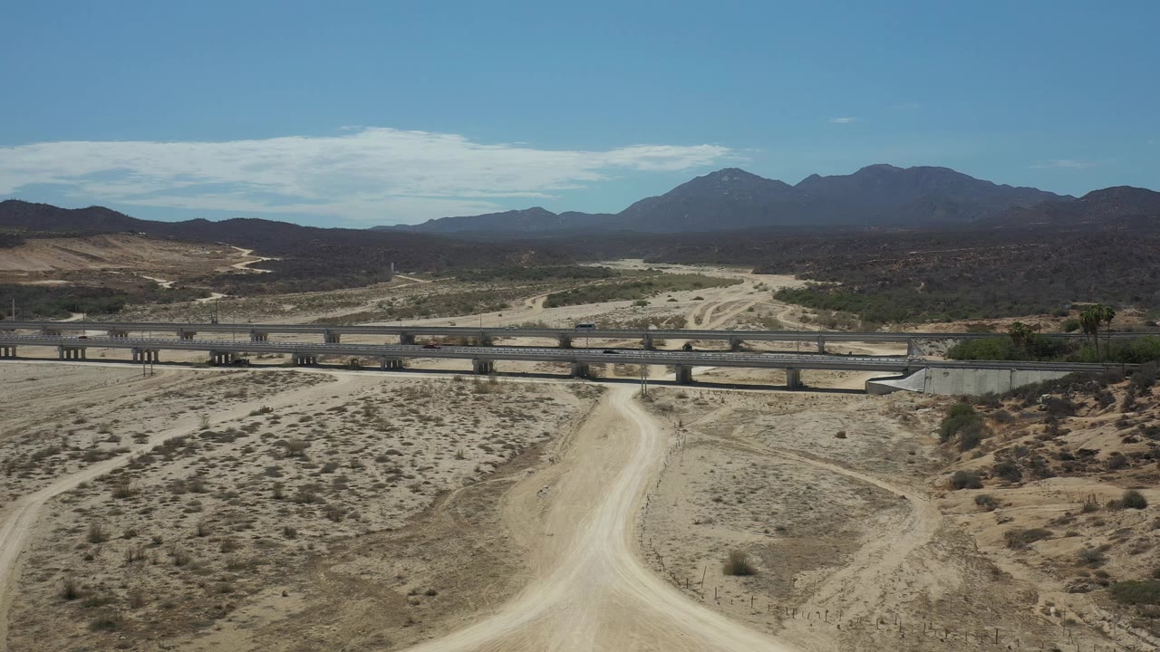 High aerial view in Los Cabos with the dirt road centered below, a bridge stretching across the frame, small vehicles on the highway, mountains beyond, and no visible people.