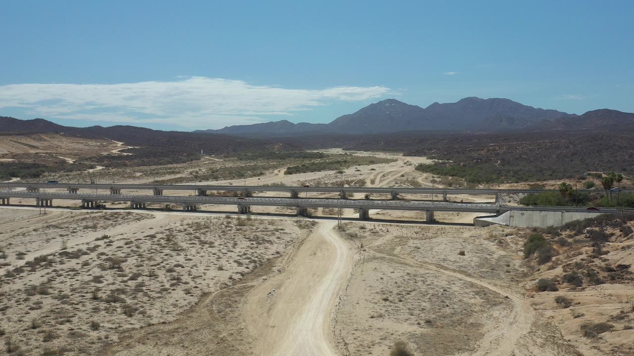 Opening frame of Los Cabos, Mexico, with a pale dirt road in the foreground, a bridge with small cars in the midground, mountains on the horizon, and no visible people.