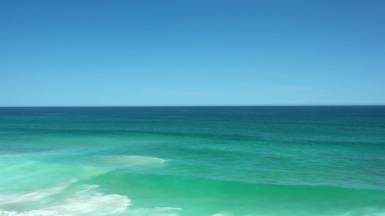 Wide Los Cabos, Mexico ocean view with a level horizon, blue sky, turquoise water, white surf at the bottom edge, and no visible people.