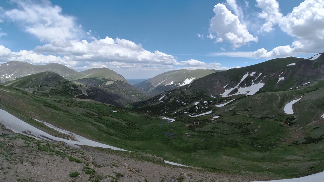 Elevated Denver Colorado mountain view with pale rocky ground at the bottom edge, green slopes through the center, broken snowfields, and no visible people.