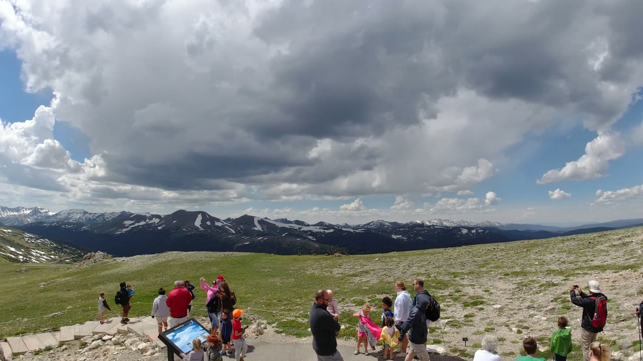 Visitors gather at a Denver, Colorado mountain overlook with an interpretive sign in the foreground and snow-streaked peaks under heavy clouds in the distance.
