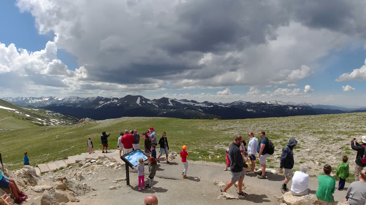 A mixed group of adults and children stands near a blue-topped overlook sign in Denver, Colorado, with rocky ground and a long mountain ridge beyond them.