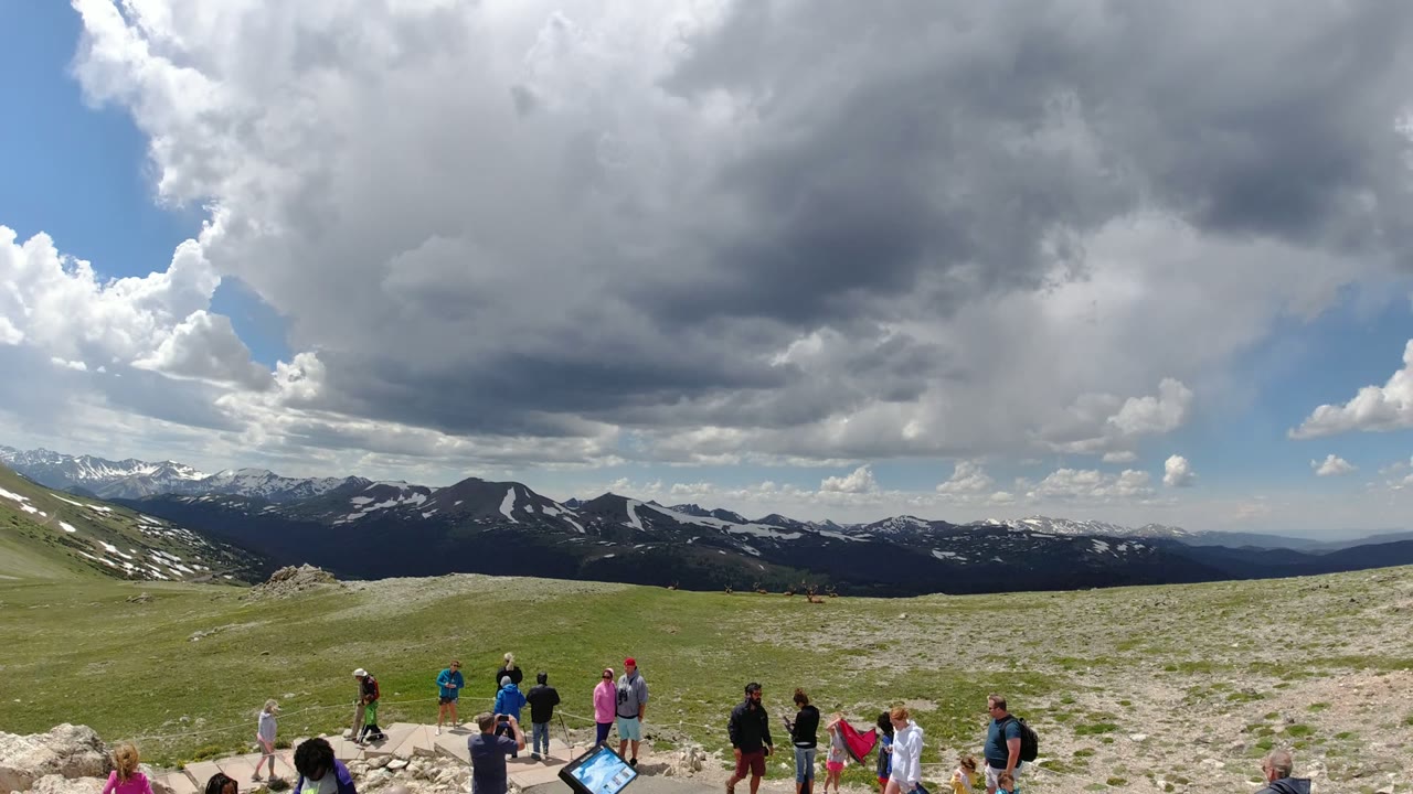 Small groups of people face the mountain range from a rocky plateau in Denver Colorado, with a signboard near the overlook and heavy cloud cover above.