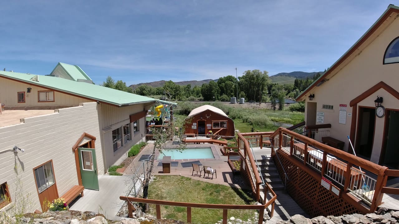 Opening frame of a Denver, Colorado exterior courtyard with beige buildings, green roofs, wood railings, a small pool, patio chairs, trees, distant hills, and no visible people.