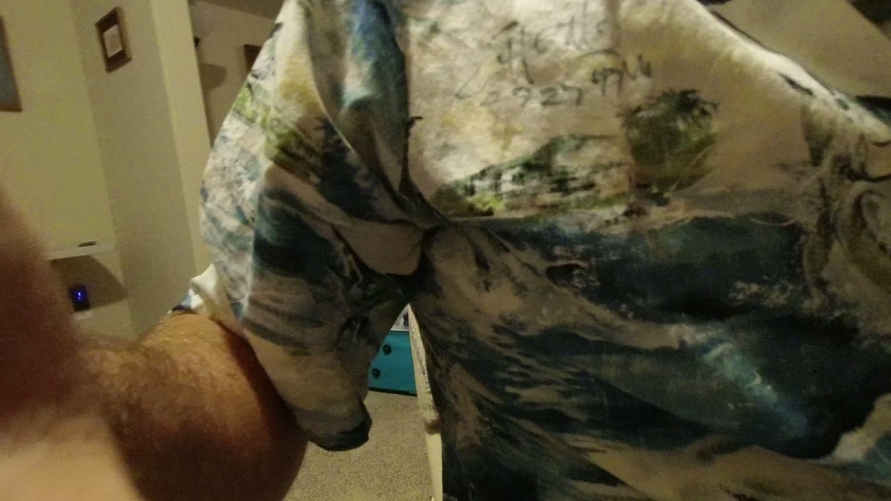 At 0:01, the camera is close to the man's patterned shirt and arm, with a finger along the right edge and beige interior walls behind him.