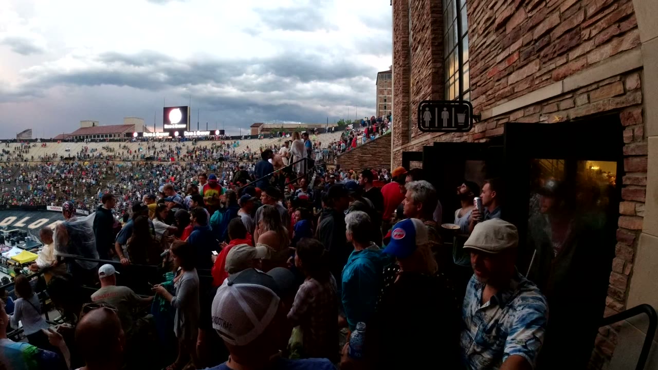 Crowded arena seating seen from a shaded concourse beside a brick wall.