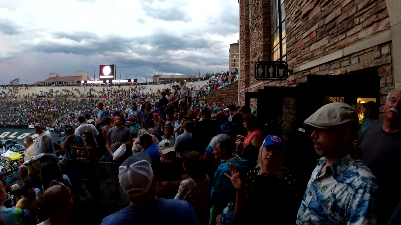 At 0:01, the Denver arena crowd remains dense while people pass through the shaded foreground aisle near the brick wall and lit doorway.