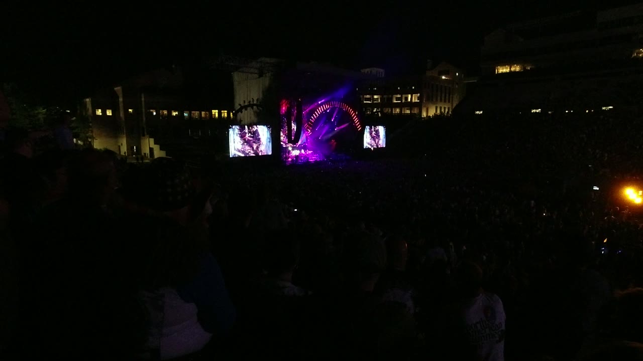 Dark audience silhouettes face a distant purple and magenta concert stage in Denver, Colorado, with two bright stage screens and lit windows behind it.