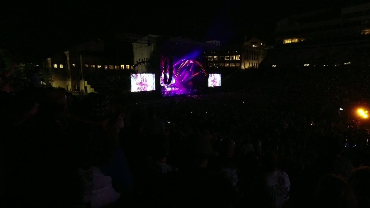Audience silhouettes dominate the lower frame as the Denver concert stage glows purple, with an orange light source visible at the right edge.