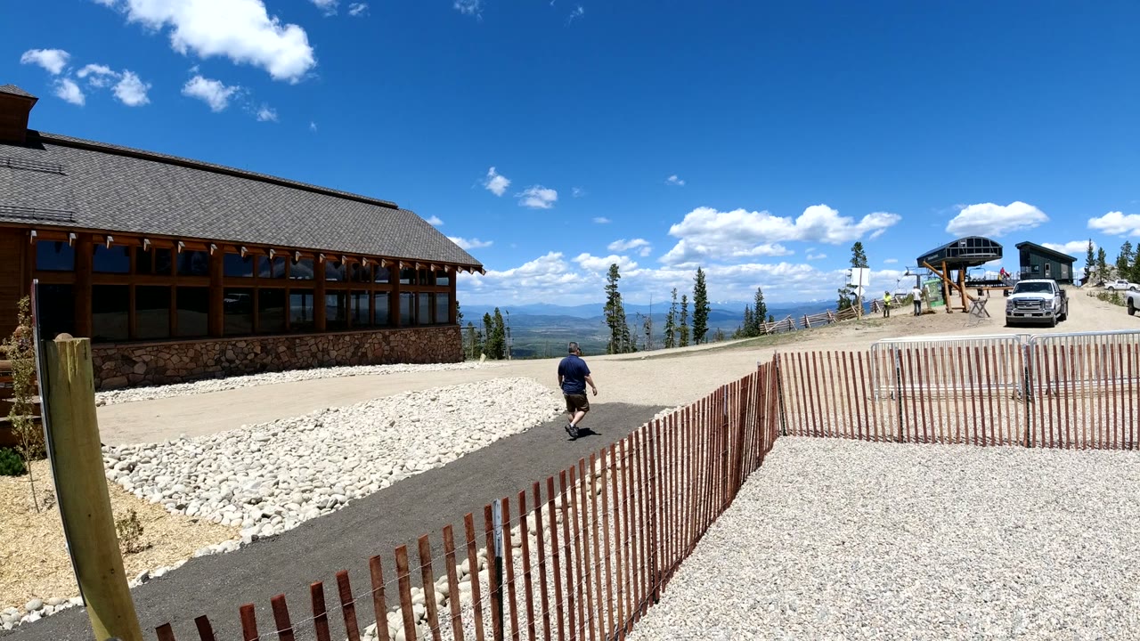 A person walks along the paved path in the Denver, Colorado mountain overlook scene, with the lodge on the left and vehicles near the right background.