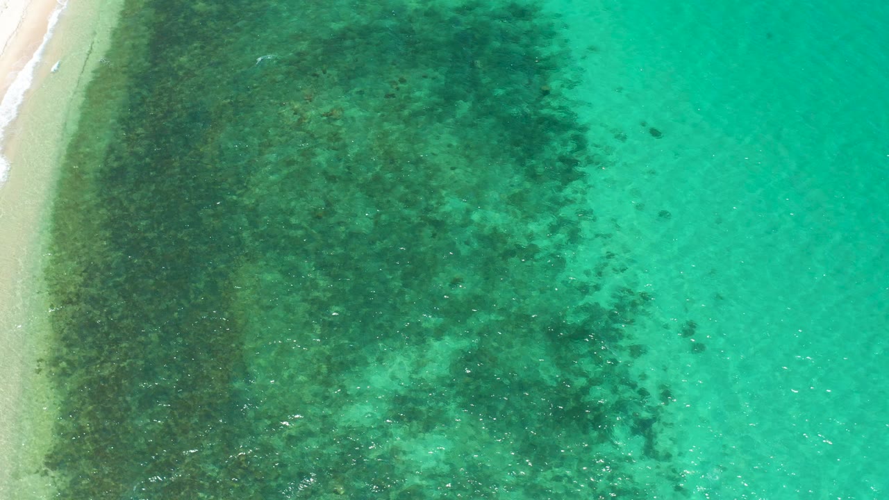 High overhead view of the El Sargento La Ventana shoreline, with pale beach at left, dark underwater patches, turquoise water to the right, and no visible people.