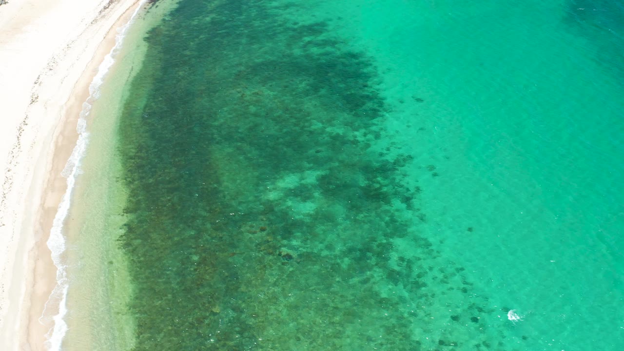 Top-down coastal view at El Sargento La Ventana with sand along the left margin, dark submerged patches across the shallows, and no visible people.