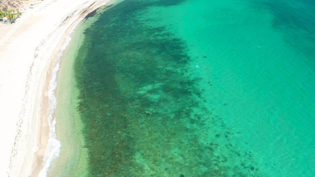 Aerial frame from El Sargento La Ventana, Mexico, showing white sand along the left edge, clear turquoise water to the right, dark underwater patches near shore, and no visible people.